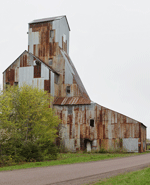 A tall rusty aluminum industrial building looms before a cloudy sky