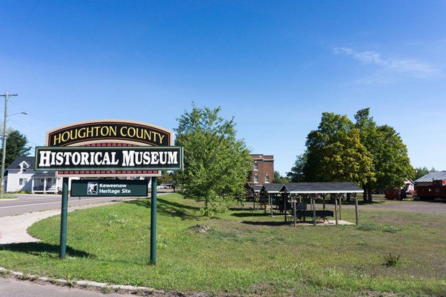 A sign along the road reading "Houghton County Historical Society," with artifacts and buildings behind it.