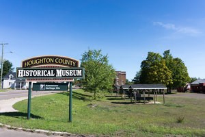 A sign along the road reading "Houghton County Historical Museum," with artifacts and buildings behind it.
