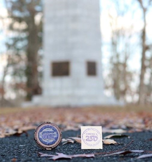 A stamp and a piece of paper with a Kings Mountain 250 stamp are on the ground with a blurred monument behind it.