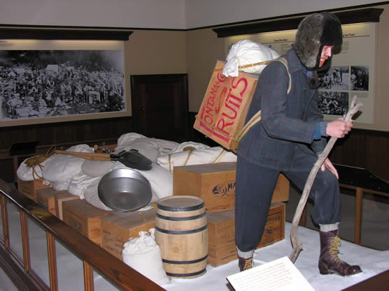 Ton of goods display showing a mannequin dressed in winter clothing of 1890s, carrying a box strapped to his back and many bags and boxes behind him