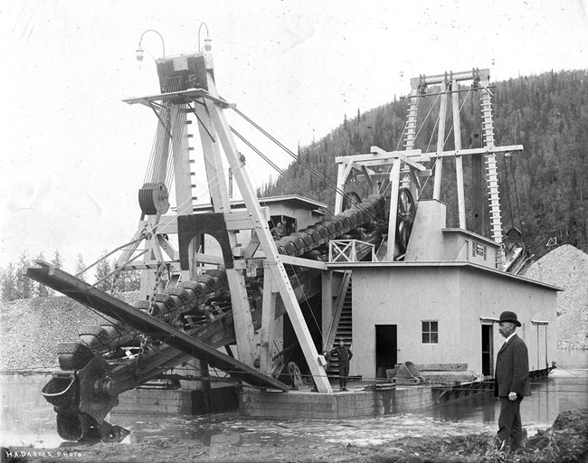 A large gold dredge with a man in a bowler hat in front of the many dredge buckets.