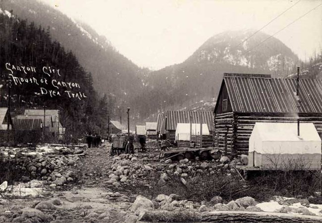 Black and white image of log buildings and tents lining a rough path in a small valley.