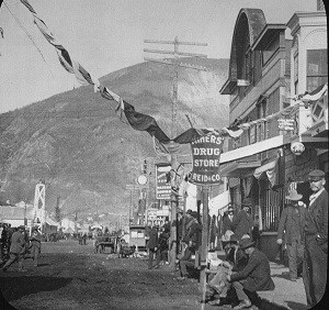 Black and white photo looking down an unpaved street lined with buildings and a mountain in the background.