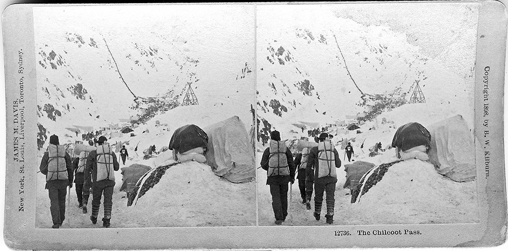Black and white stereoscope image of hikers with packs walking away from the camera toward a narrow valley with people climbing a snowy pass in background.