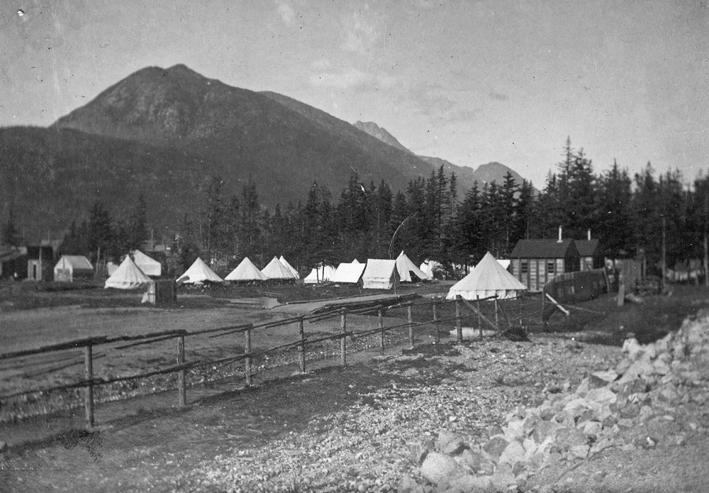White tents in cleared area with cabins, trees, and mountain backdrop.