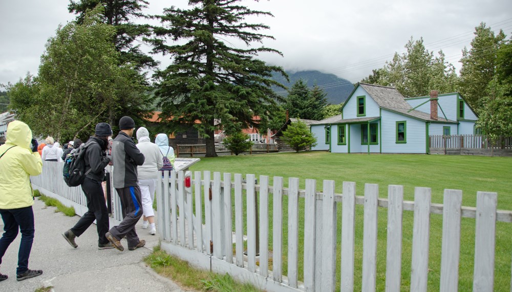 People approach a blue house with a green lawn and picket fence. People approach a blue house with a green lawn and picket fence.