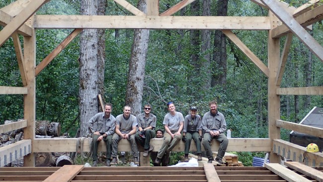 Group of six people sit on an exposed beam.  A man stands below the middle of the group.