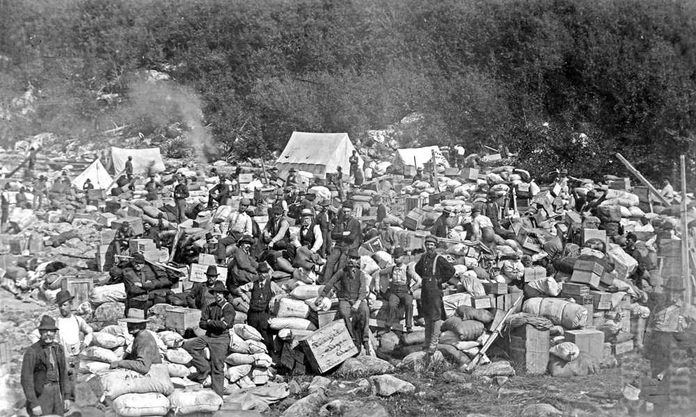 Black and white photo of men posed with piles of boxes and sacks in the foreground and white canvas tents in the back.