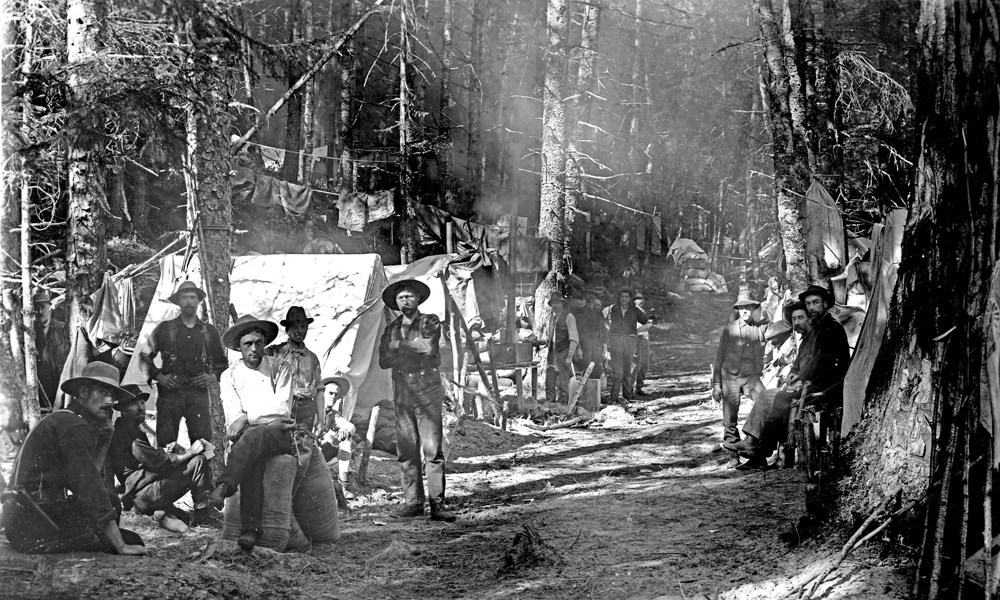 Men sit at a tent camp along a forested trail.