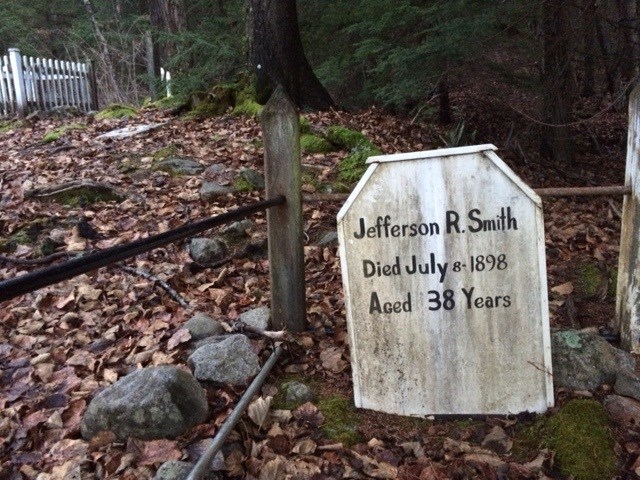 Wooden grave marker with writing "Jefferson R. Smith Died July 8 - 1898 Aged 38 Years" inside a fence with leaves on the ground.