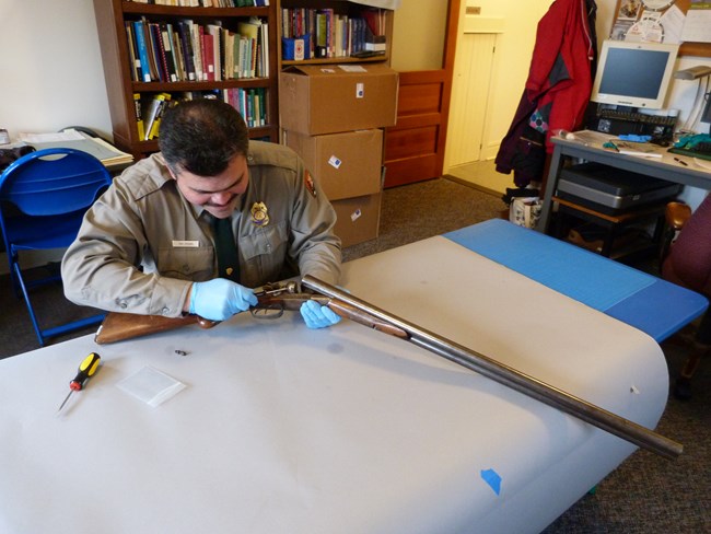 color photo of park ranger wearing blue protective gloves seated at a long blue table examining a shotgun