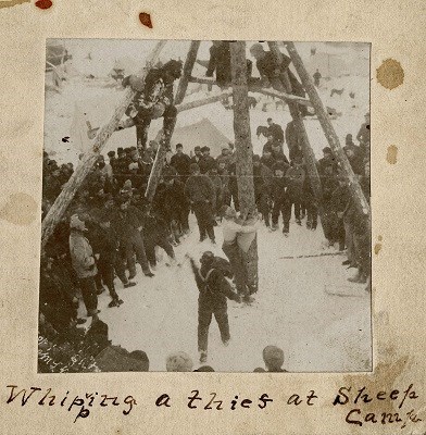 Black and white photograph of a crowd watching a man tied to a pole.  Text reads "Whipping a thief at Sheep Camp"