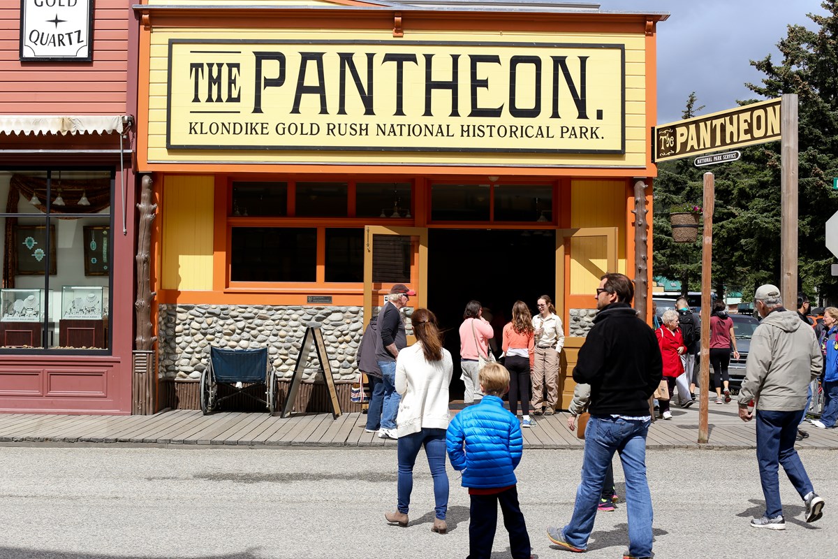 People walk in front of a brightly colored building with sign "Pantheon Saloon" People walk in front of a brightly colored building with sign "Pantheon Saloon"