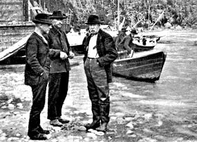 Black and white photo of three men standing at the edge of water with boats behind them. Black and white photo of three men standing at the edge of water with boats behind them.
