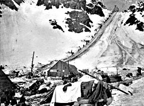 Black and white photo of buildings in foreground and line of people headed up a mountain pass. Black and white photo of buildings in foreground and line of people headed up a mountain pass.