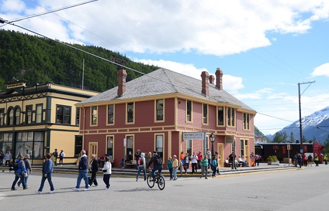 People cross streets at an intersection in front of a large, red building. People cross streets at an intersection in front of a large, red building.