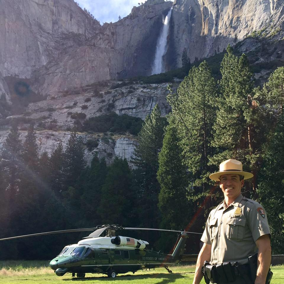 Jordan Neumann a ranger poses in front of a helicopter and waterfall