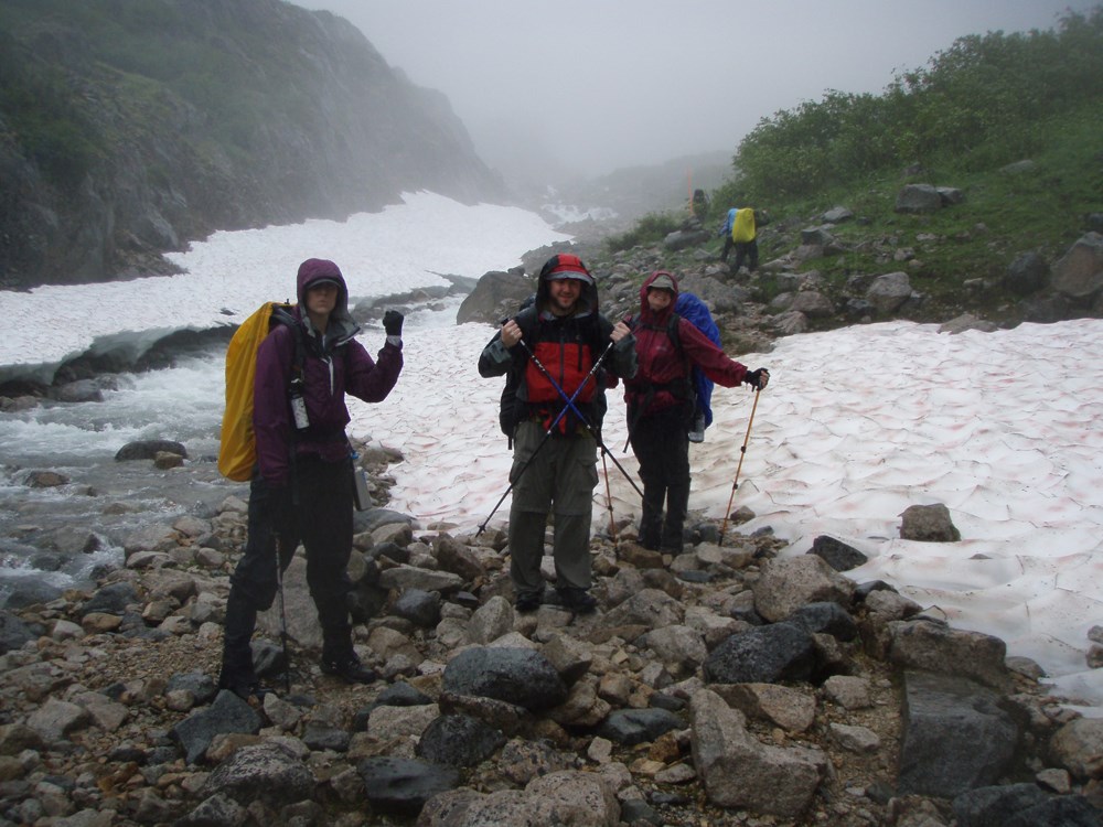 Three hikers pose for the camera in rain gear while standing on a rocky area surrounded by snow and fog.