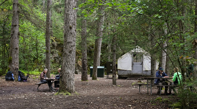 People sit at picnic tables in brush free area in front of a white tent structure