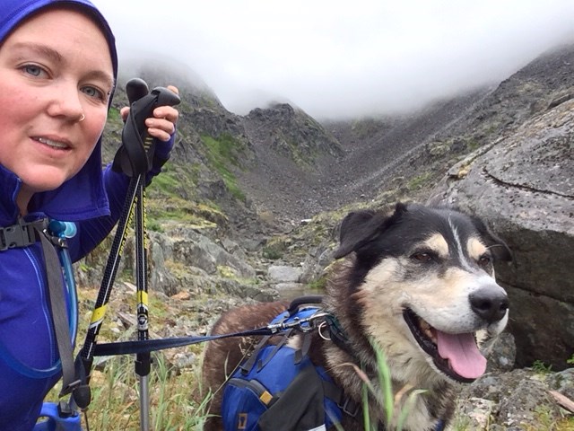A color selfie style photo of a woman in a raincoat with a leashed dog wearing a backpack.