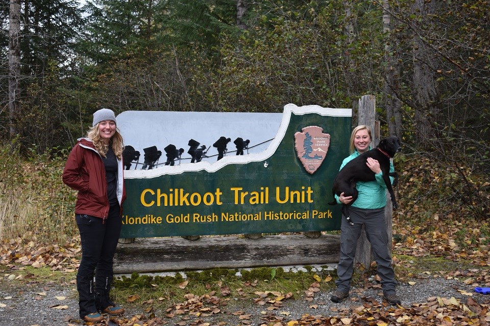 Two hikers and a dog being held by one of the hikers pose in front of a sign that says "Chilkoot Trail Unit".
