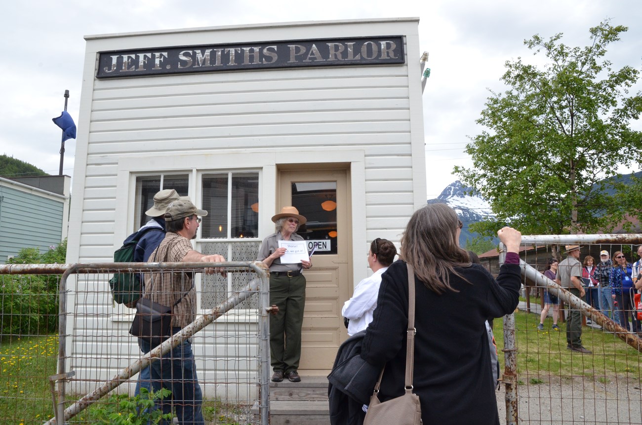 Facing the camera, a ranger stands on the steps of a small building with a sign "Jeff. Smiths Parlor" while people face her
