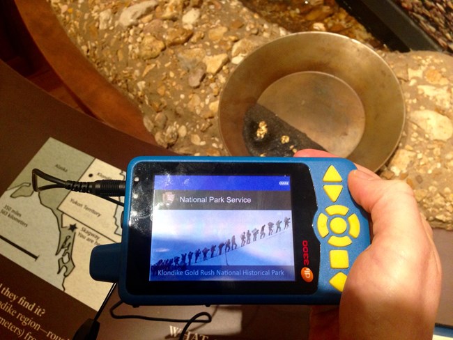 Visitor holds a Durateq audio description device by a map and model gold pan exhibit