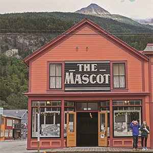 Two people taking pictures in front of a building titled "The Mascot"  with a mountain peak in the background