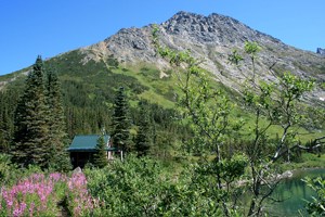 Cabin and vegetation a long a clear lake with mountain background.