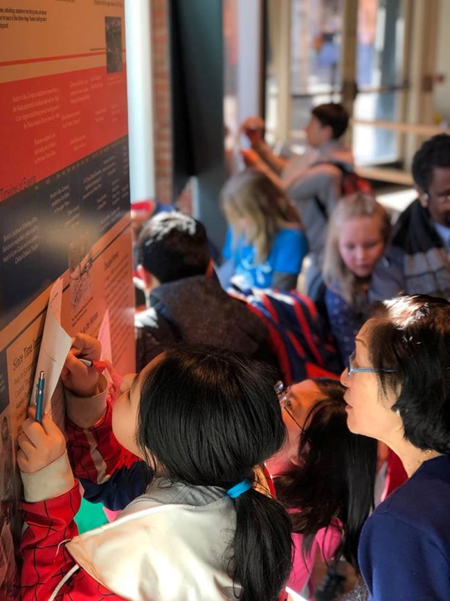 children look at exhibits and write on paper while an adult stands by