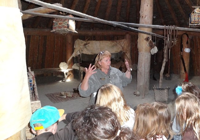 Female adult speaks in front of students inside the earthlodge.