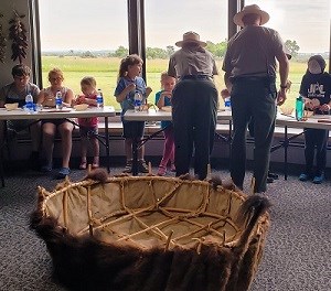 6 kids sit at tables inside working on craft. 2 rangers stand and support them. Large bull boat in foreground.