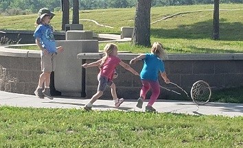 1 girl jumps high outside as 2 other girls throw their sticks towards a rolling hoop.