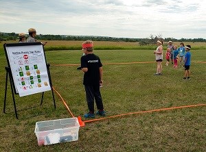 7 kids stand on grass within an orange square boundary. 2 rangers stand behind a Native Prairie Tag Rules poster.