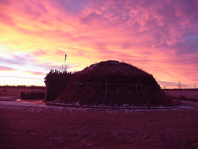Clouds of white, yellow, pink, and purple overhead an earthlodge.