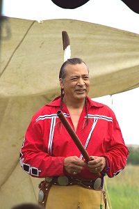 Keith Bear Man with braids and a feather tucked vertically in his hair holds a wooden flute outside..
