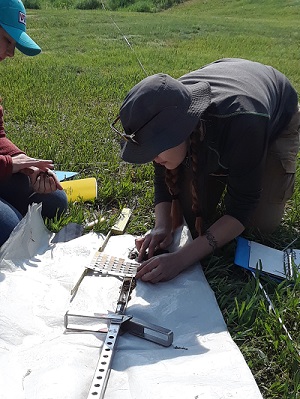 Matching Soil Samples Female student leans over a tarp outside and looks at soil in a tube and a color information card.
