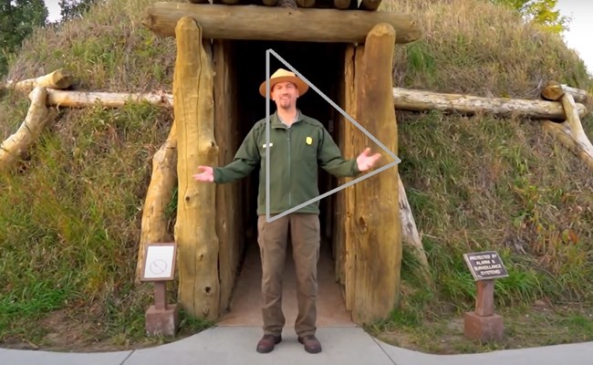 A ranger stands in front of an earthlodge with a play button used for videos in front.