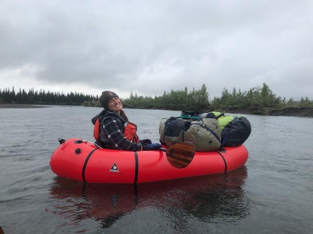 Girl on red raft floating on a river with NPS cap and paddle across lap. River is calm and trees line the background. 