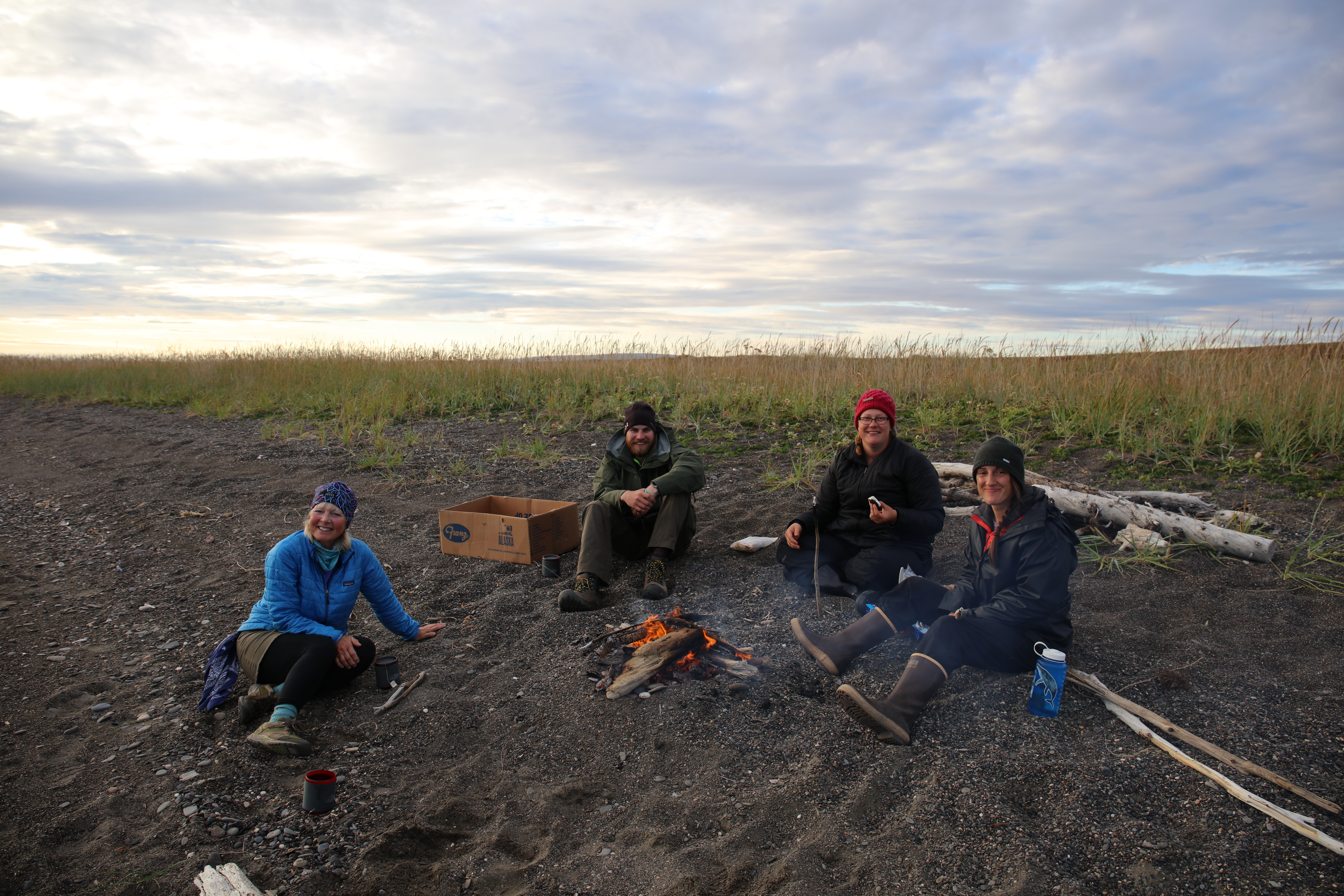 Three females and one male sit on the gravel beach next to a burning campfire.
