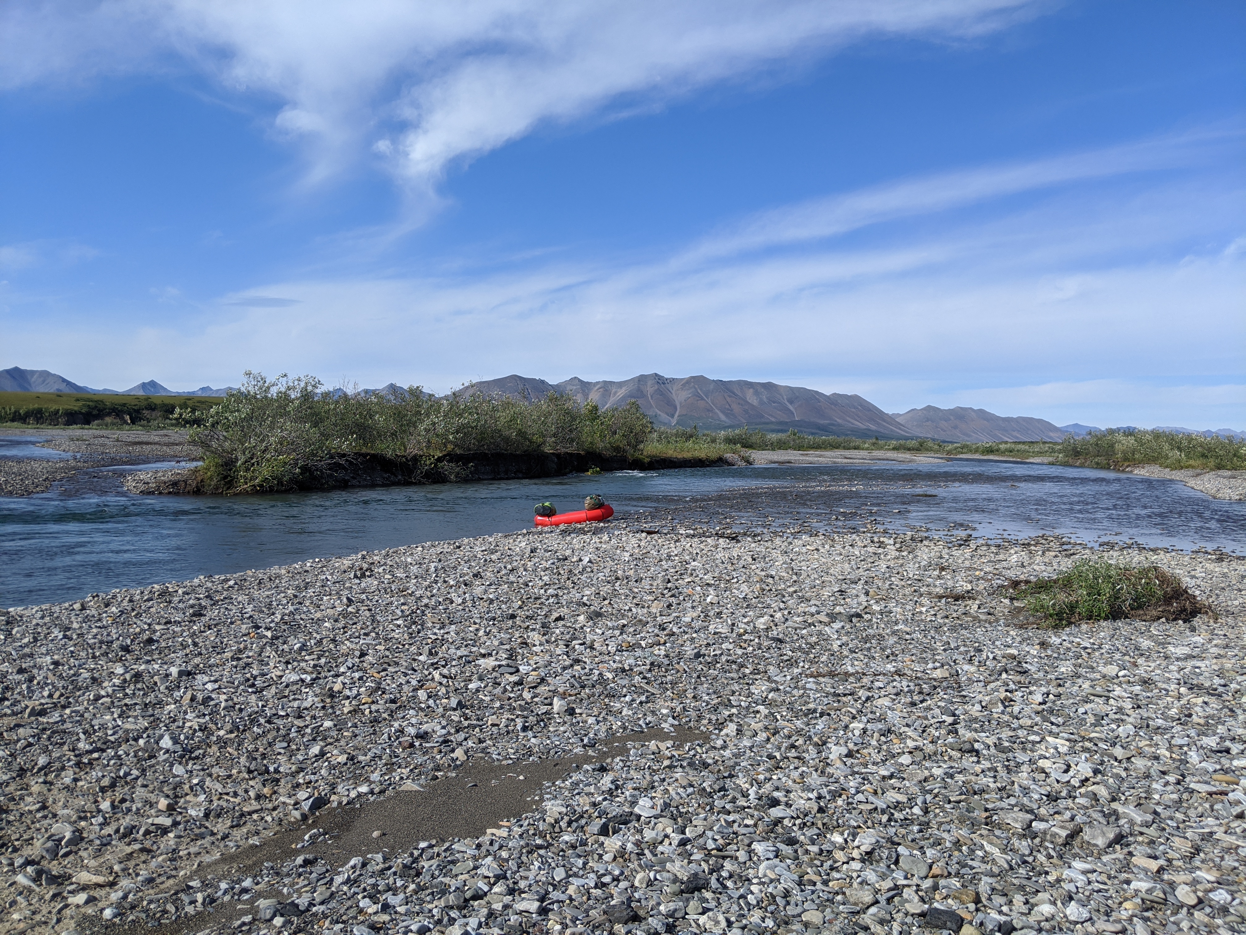 A red raft rests on a gravel bar on the side of a river with a view of mountains, trees, and blue sky with clouds in the background. 