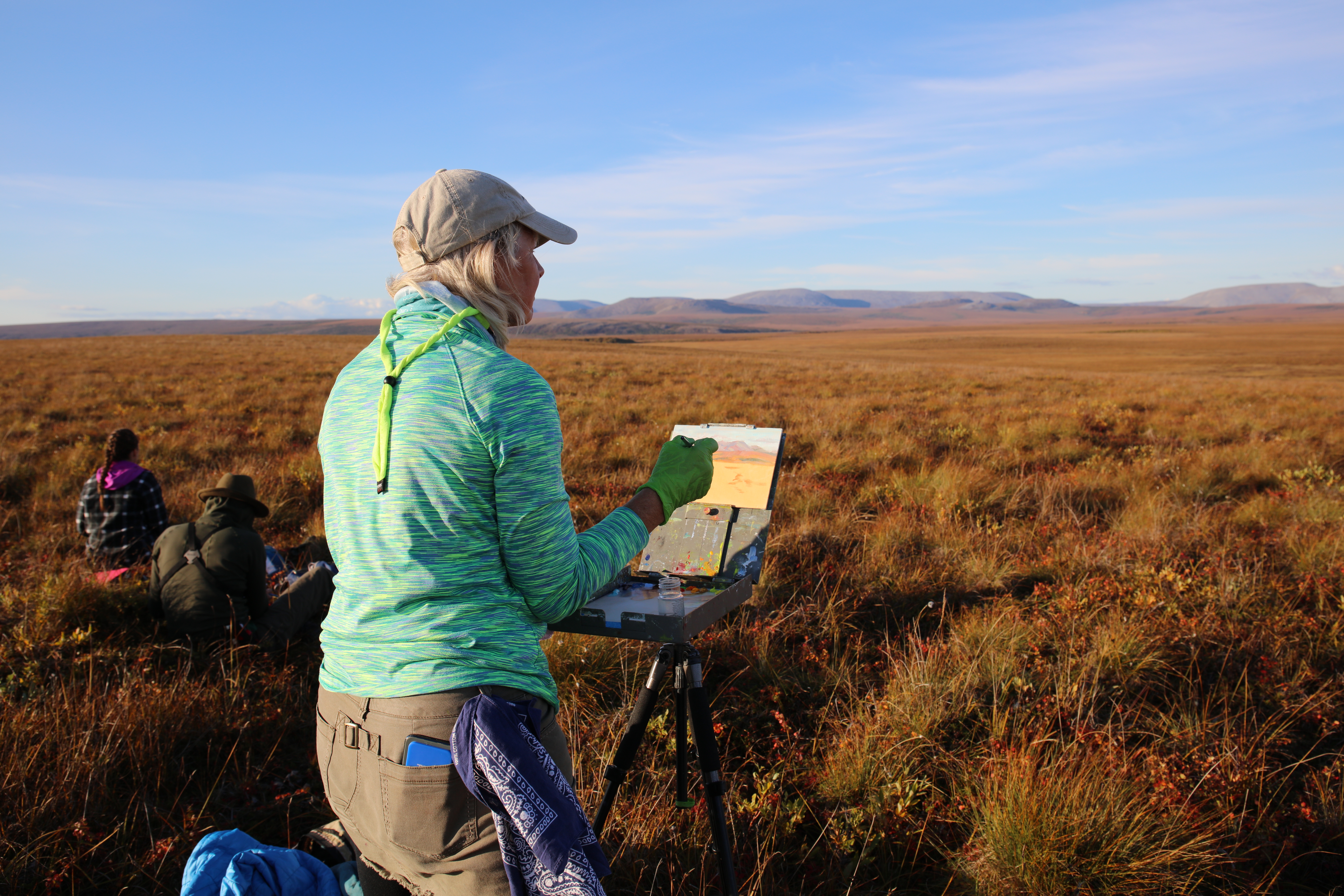A woman paints in a field of tundra while two park rangers sketch the mountains. 