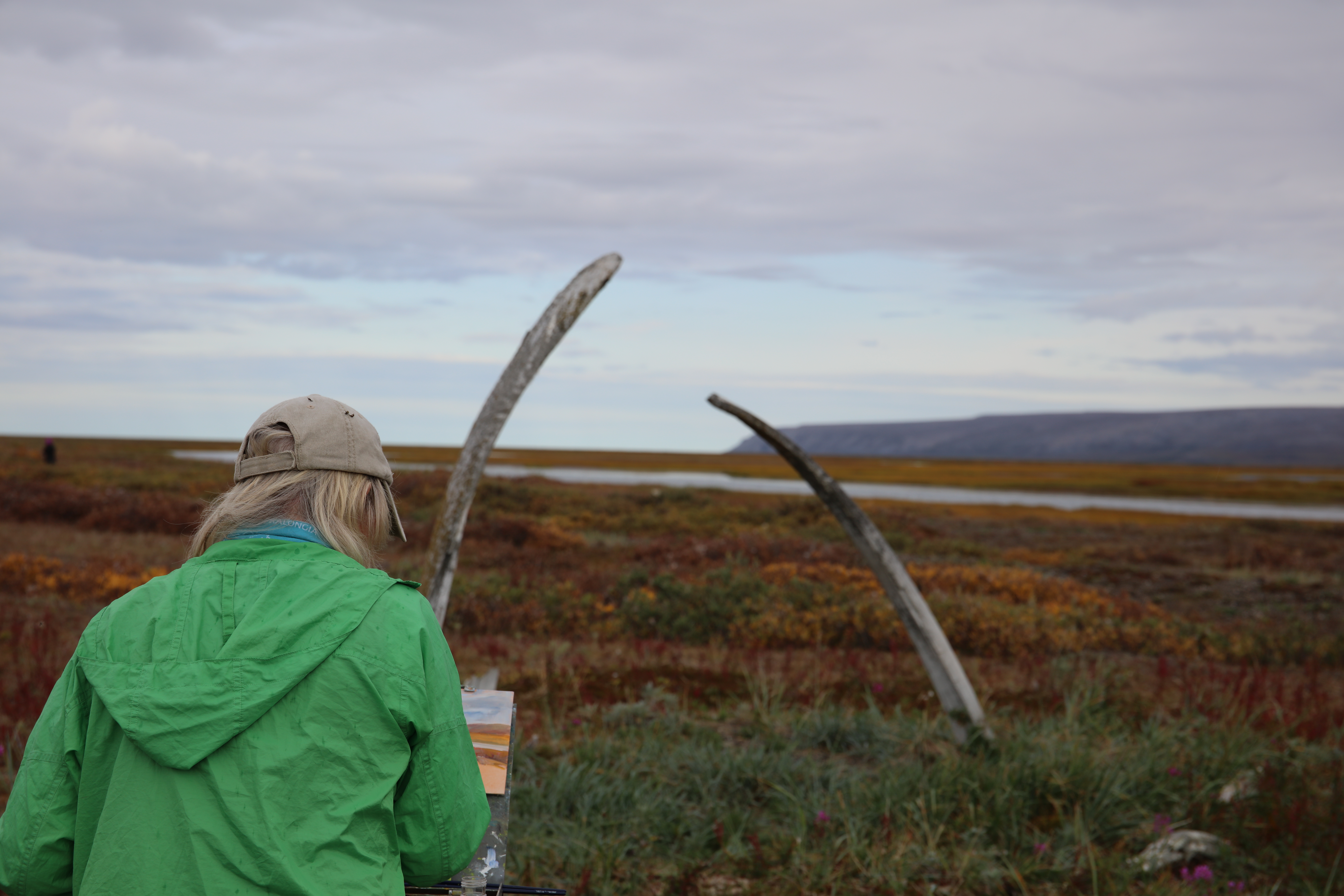 A woman paints the scene of whale jaw bones and tundra in front of her. 