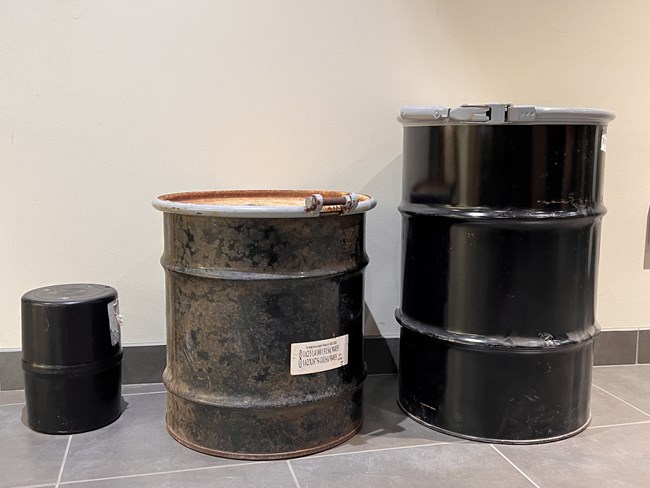 Image of three black barrels in varying sizes lined up from smallest to largest in front of a tan wall and on a grey floor.