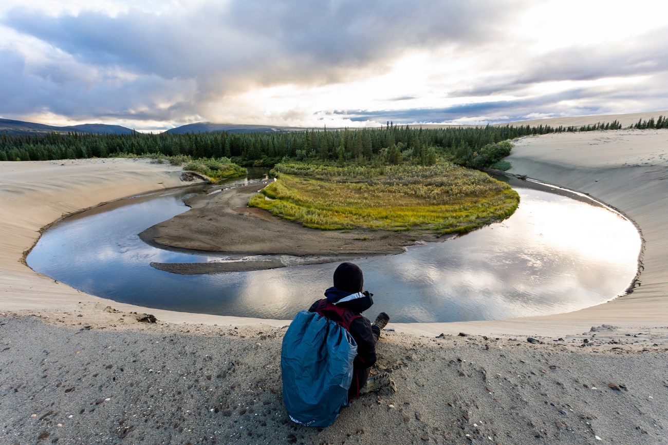 Hiker stops near Niaktovik Creek. Sand dunes with creek and forest in background.