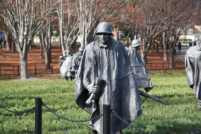 A stainless-steel statue stands in a field of green with a helmet and weapon.
