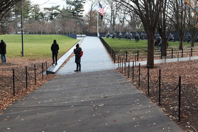 The pathway to the memorial transitions from sidewalk to granite concrete as visitors approach the memorial. Stain-less steel statues of veterans stand to the right.