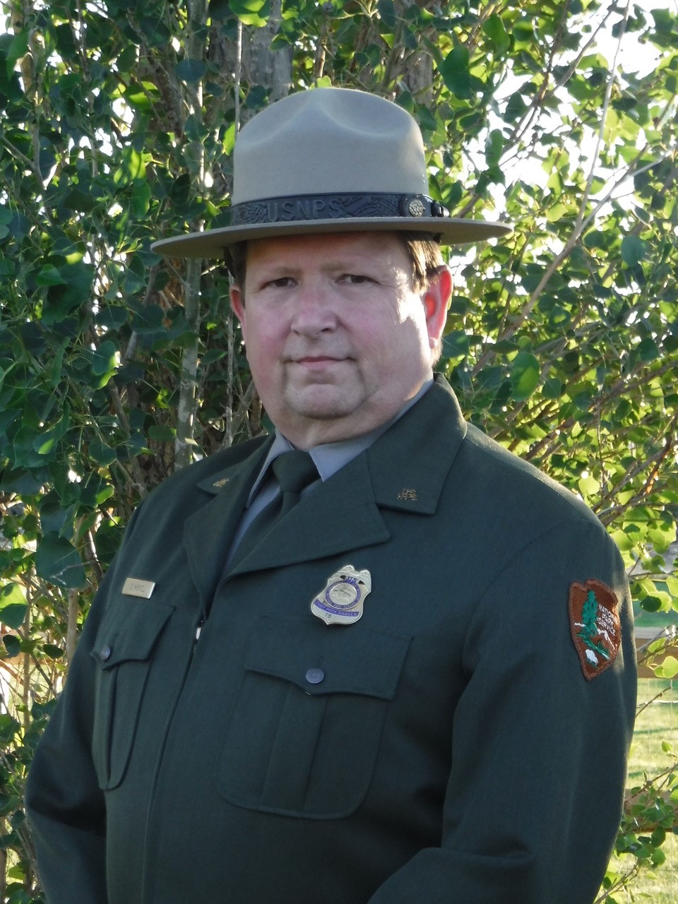 blobid3_1627577641524 A head-and-shoulders portrait of Chris Mengel in full NPS formal uniform and flat hat, looking at the camera with a serious expression with green foliage in the background.