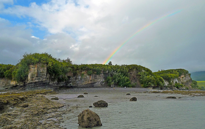 a beach with forested ridge line and a rainbow arcing overhead